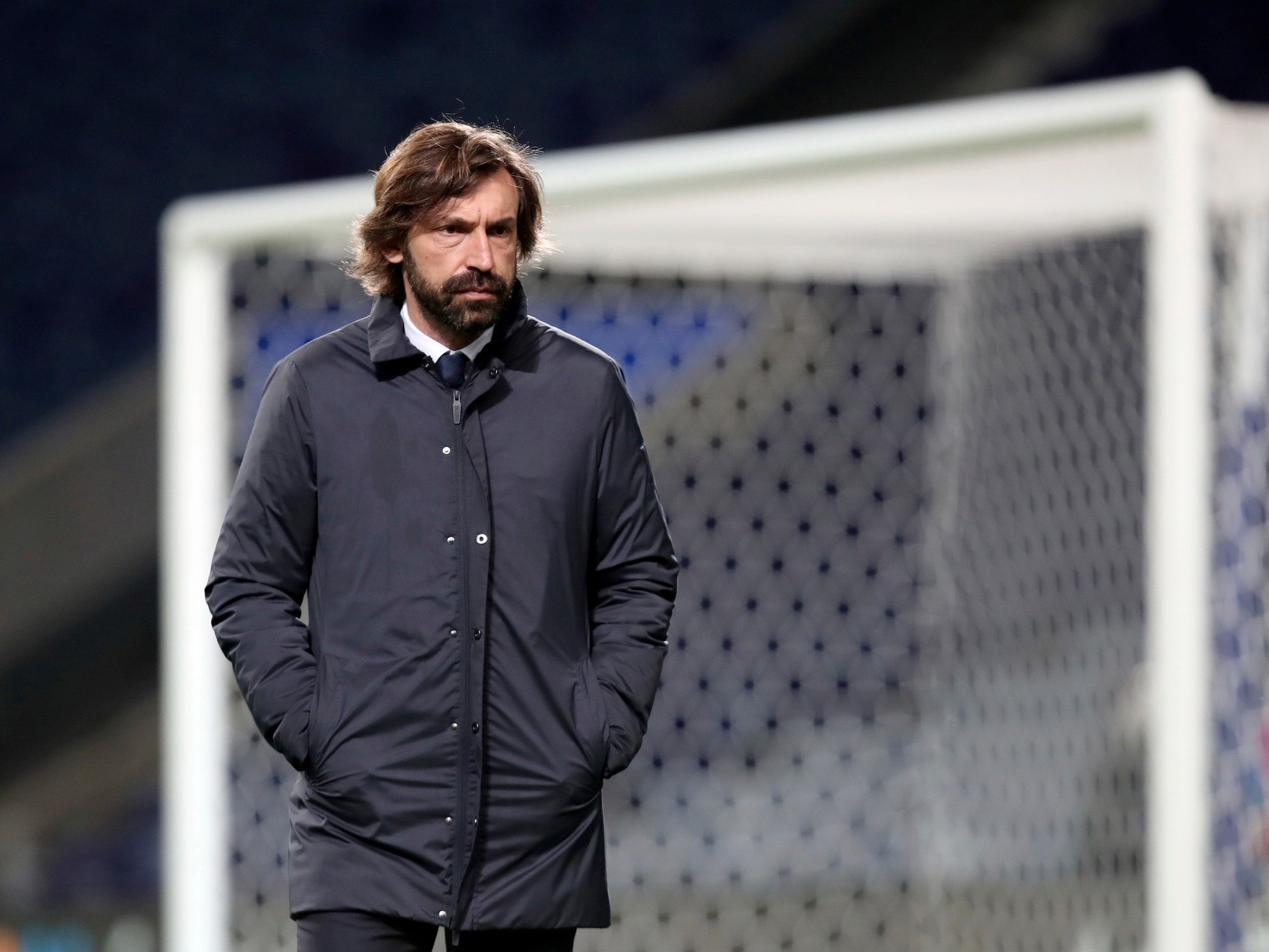 Andrea Pirlo, de campera oscura y con las manos en los bolsillos, camina por el costado del campo frente a un arco durante un partido