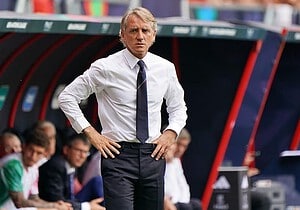 Roberto Mancini de camisa blanca y corbata oscura, con las manos en la cintura, parado al costado del campo frente al banco de suplentes