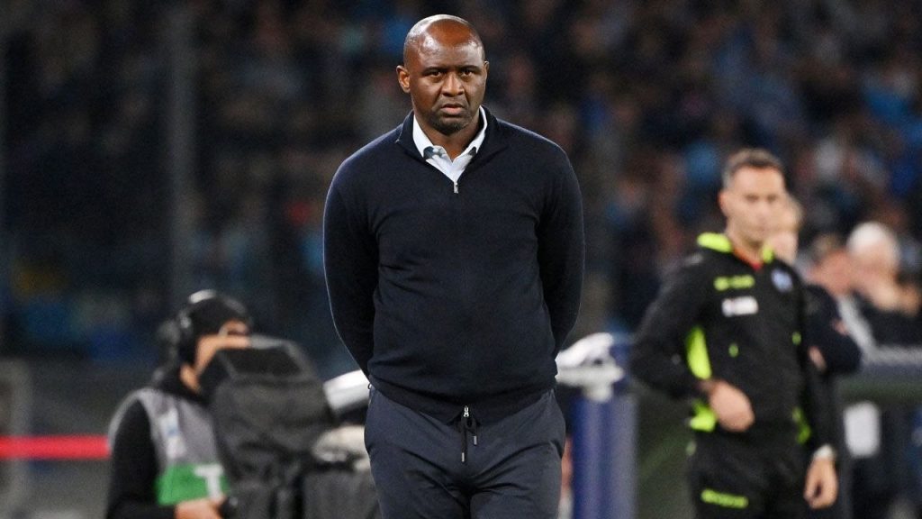 Patrick Vieira camina por el costado del campo con buzo azul oscuro, de espaldas al banco de suplentes, durante un partido nocturno