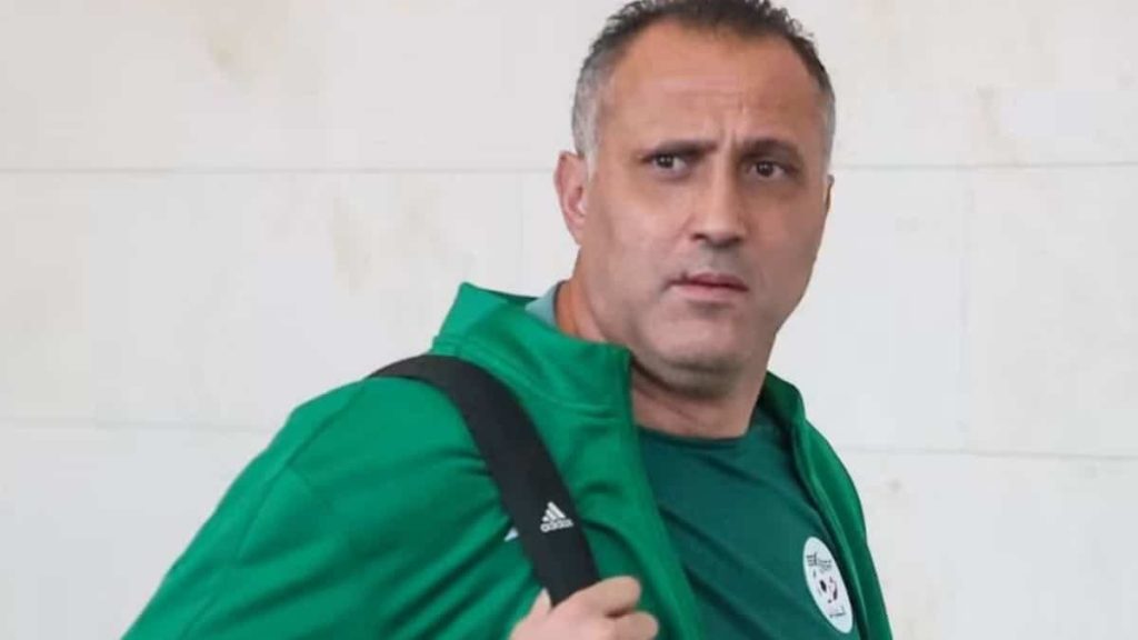 Brahim Hemdani con la campera verde de la selección argelina y un bolso al hombro, mirando a cámara frente a una pared blanca
