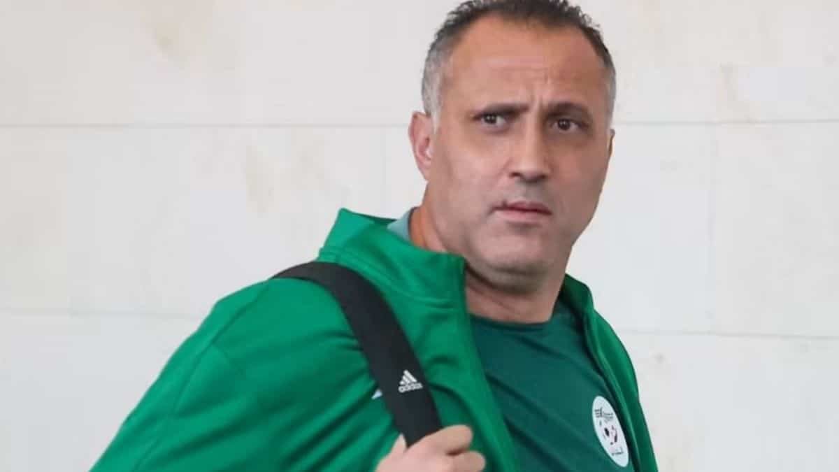Brahim Hemdani con la campera verde de la selección argelina y un bolso al hombro, mirando a cámara frente a una pared blanca