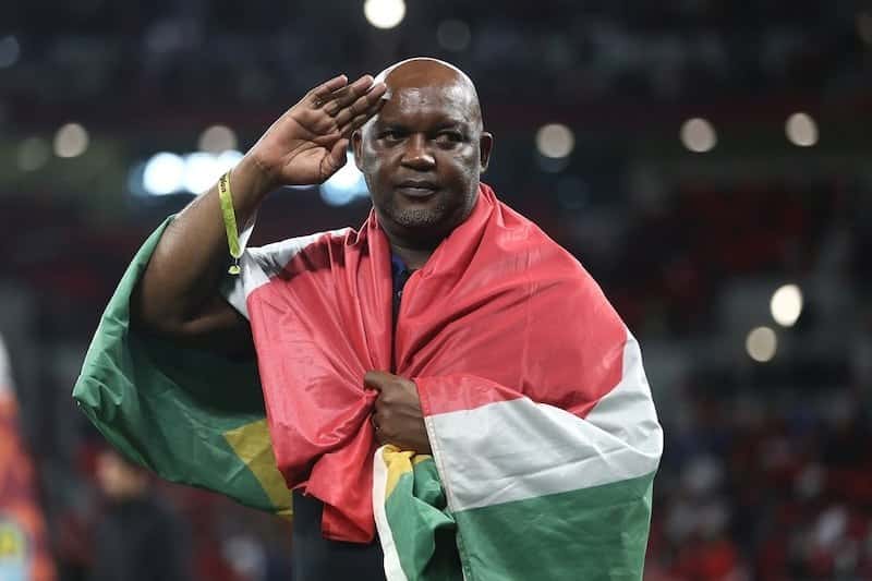 Pitso Mosimane saluda con la mano en la frente, envuelto en una bandera de Sudáfrica, en el campo de juego de un estadio