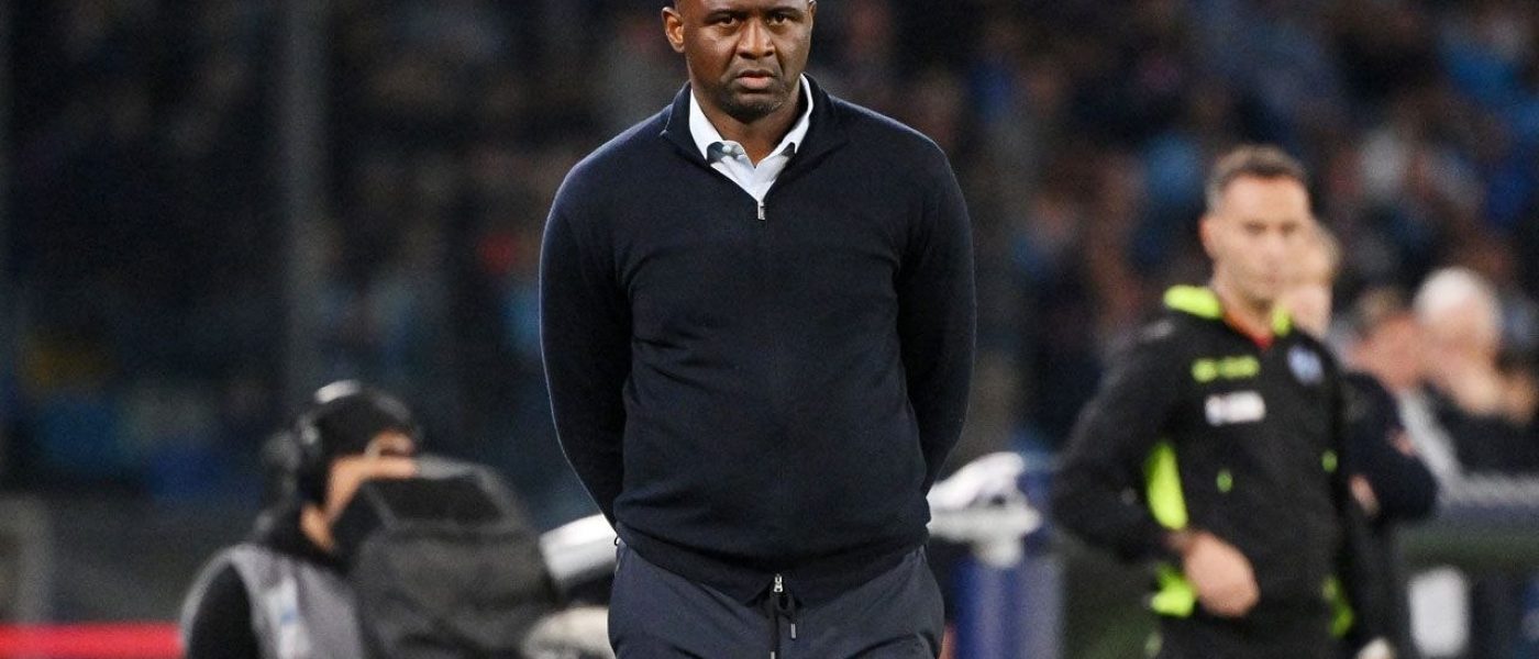 Patrick Vieira camina por el costado del campo con buzo azul oscuro, de espaldas al banco de suplentes, durante un partido nocturno