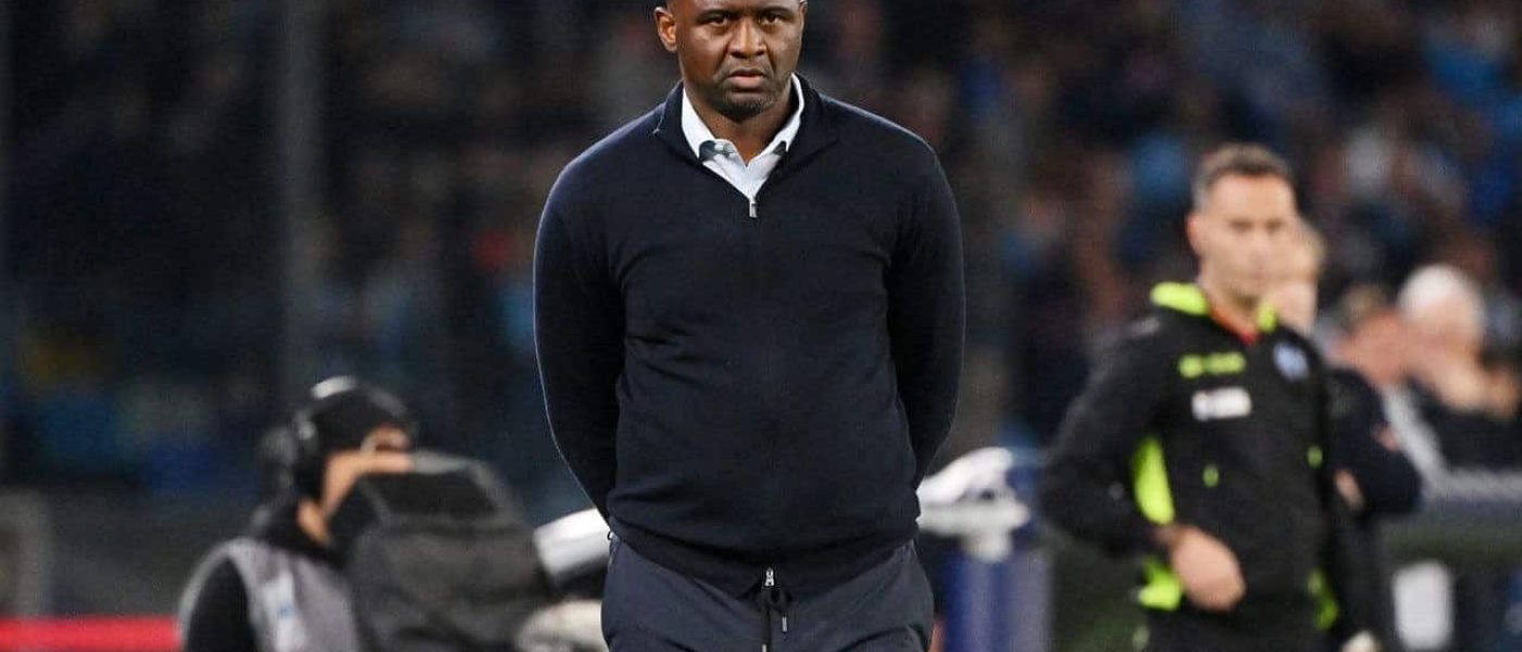 Patrick Vieira camina por el costado del campo con buzo azul oscuro, de espaldas al banco de suplentes, durante un partido nocturno