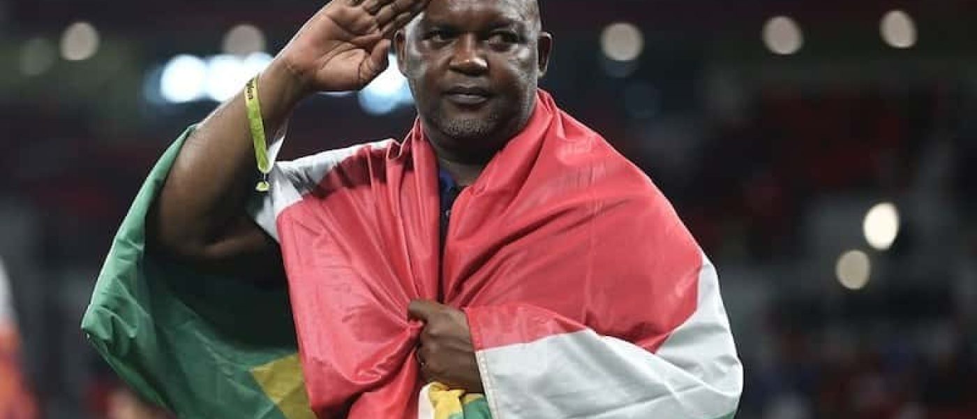 Pitso Mosimane saluda con la mano en la frente, envuelto en una bandera de Sudáfrica, en el campo de juego de un estadio