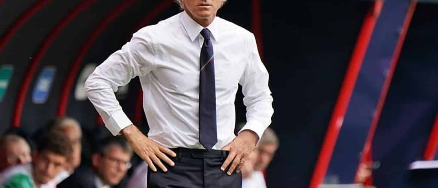 Roberto Mancini de camisa blanca y corbata oscura, con las manos en la cintura, parado al costado del campo frente al banco de suplentes