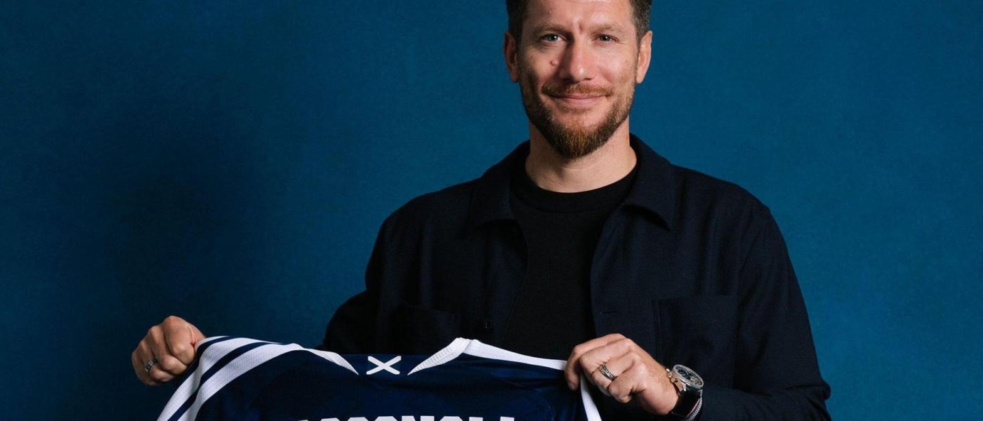 Sebastien Pocognoli sonrie mientras sostiene una camiseta azul de la seleccion de Escocia con su apellido estampado en la espalda